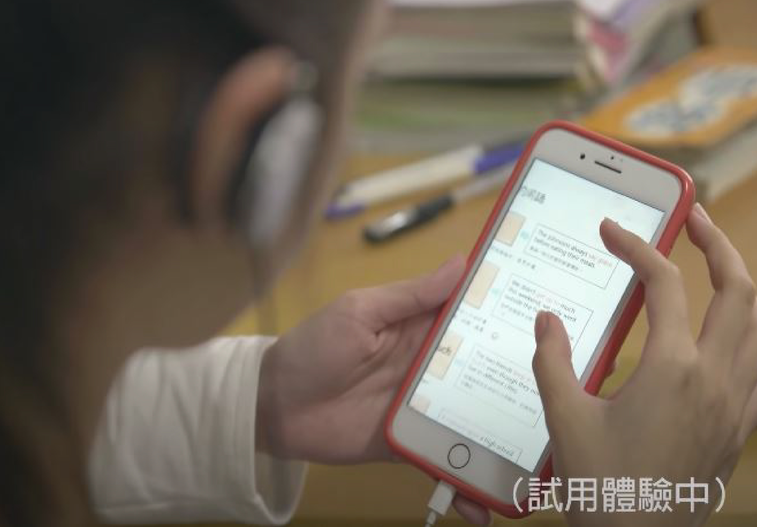 一個人手持紅色iPhone，屏幕顯示聊天對話内容。背景為桌面，放有耳機、筆和其他文具。屏幕右下角有「試用體驗中」的字樣。
