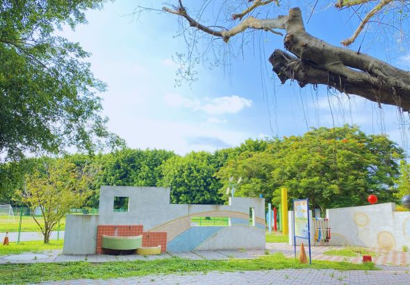 公園內設有現代感的灰色混凝土遊樂設施，包括階梯、滑梯和攀爬區。大樹枝條在上方垂落，周圍被綠色樹木和草坪包圍，晴空萬里，環境寬敞明亮，適合兒童遊憩。