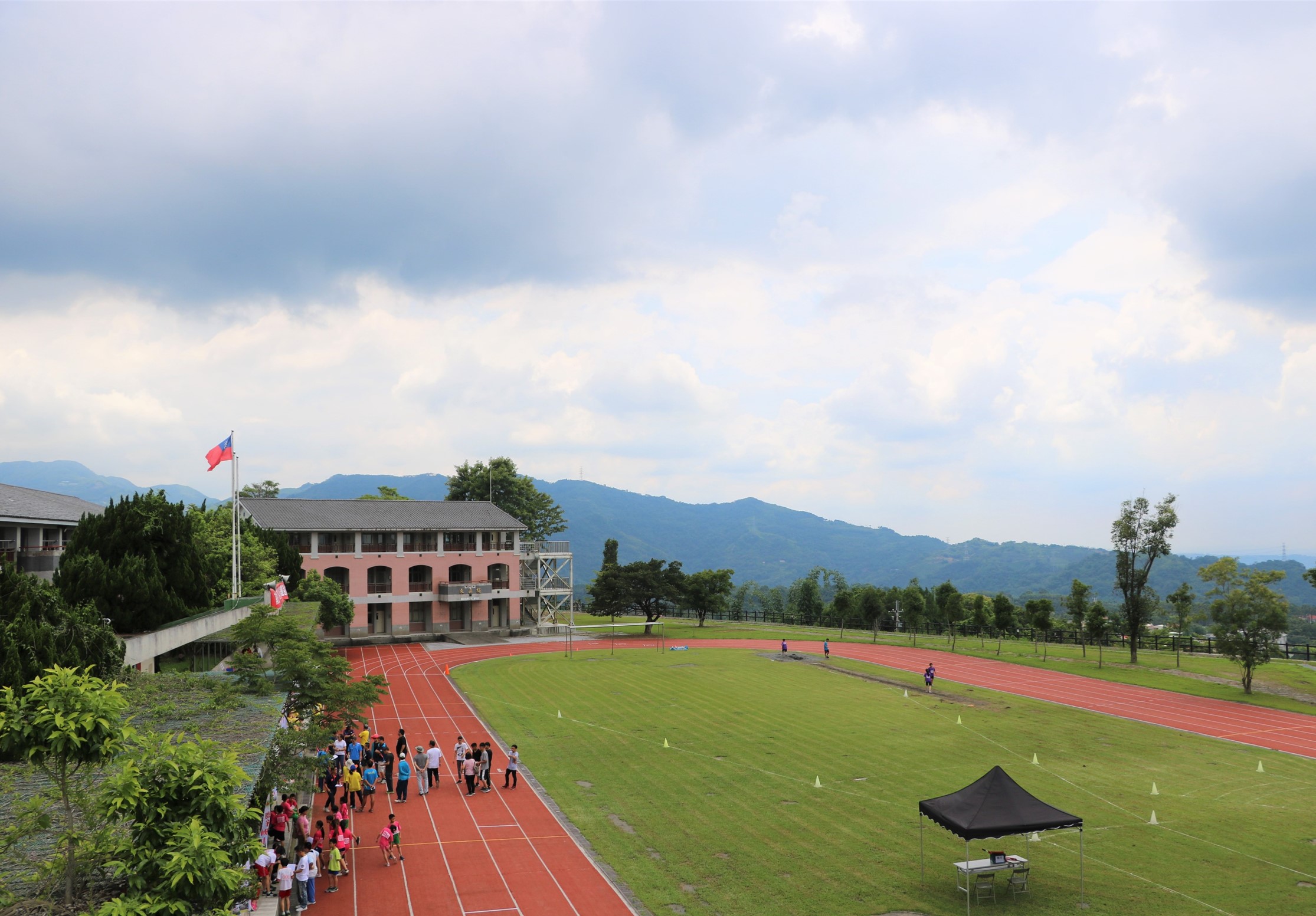 山區學校操場全景，紅色跑道上聚集多位學生進行活動，綠色草坪上設置訓練標誌，粉紅色建築物飄揚國旗，遠方群山環繞，晴朗天空。