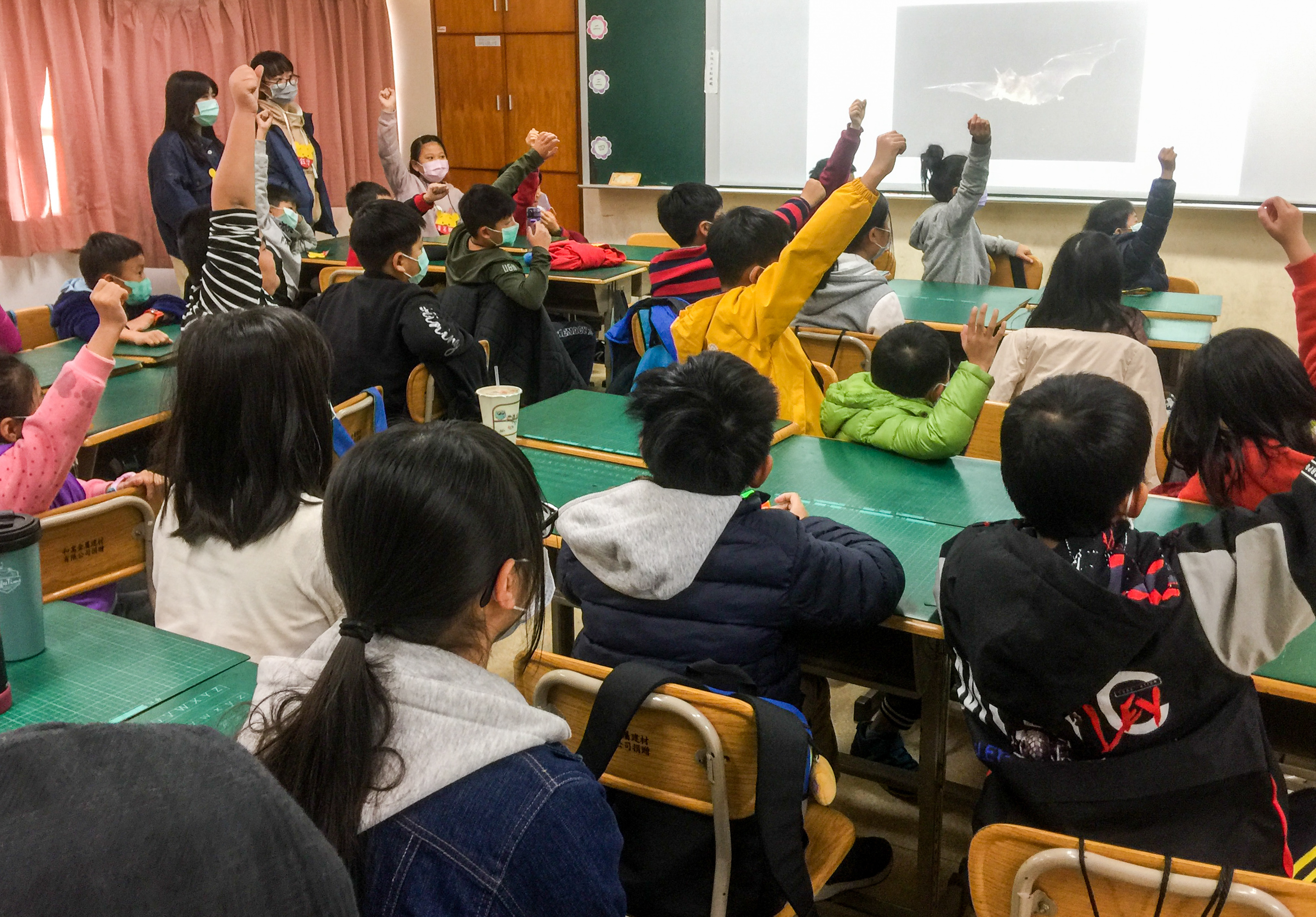 教室內舉辦海洋生物教育活動，學生們坐在綠色課桌前舉起雙手踴躍參與。講台上播放海洋生物投影片，兩位教師站在前方帶領互動式教學。學生穿著冬季服裝，戴著口罩，展現高度學習熱情。