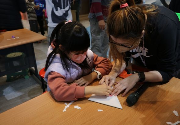 一名戴眼鏡的女童坐在木桌前，一位成人教師在她身旁指導，兩人一起進行手工藝活動，桌上散放著白色紙片碎屑。背景有其他學生和成人站立，呈現課堂或工作坊的教學場景。