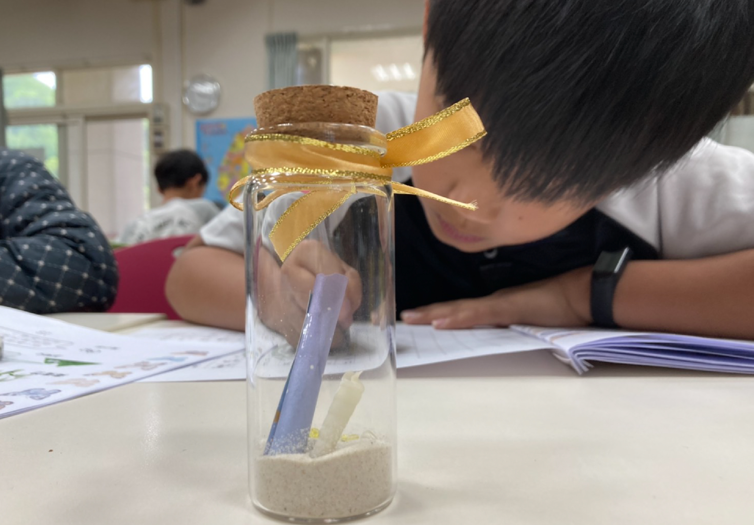 教室內，學生傾身觀察一個透明玻璃罐。罐中裝有沙子和一支藍色試管，罐口用軟木塞和金色緞帶裝飾。背景可見其他學生在進行科學實驗，桌上擺放筆記本和教材。