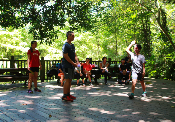 在綠樹成蔭的公園廣場上，一群年輕人進行跳繩運動。中央一位穿著黑衣的男性正在跳繩，周圍站著穿著紅白運動服的同伴觀看和參與。後方長椅上坐著數位觀眾，整個場景充滿青春活力和運動氛圍。