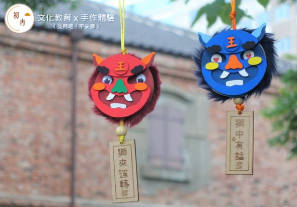 兩個手工布藝掛飾懸掛在古建築前，左側為紅色惡魔臉譜，右側為藍色怪獸臉譜，各配有木製標籤。展示文化教育與手作體驗的創意活動成果。