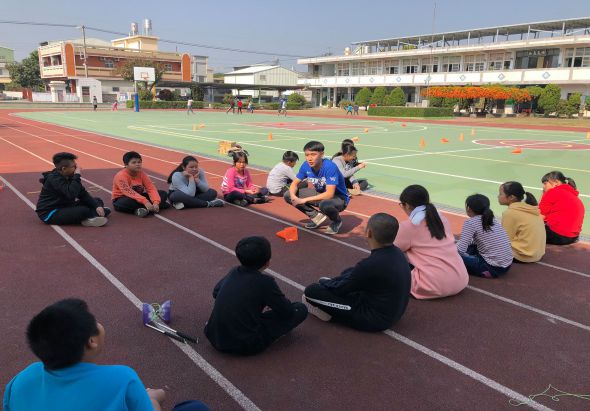 學校運動場上，一群學生圍坐成圈，進行戶外體育課程。講師在中央示範動作，背景可見校舍、綠色運動場和花卉布置。