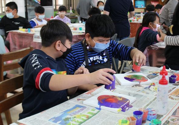 兒童藝術工作坊現場，戴著口罩的兩名小朋友坐在桌前，專注地進行手工藝創作。桌上擺放著色彩繽紛的顏料、畫筆和美勞用具，背景可見其他參與活動的兒童和家長。