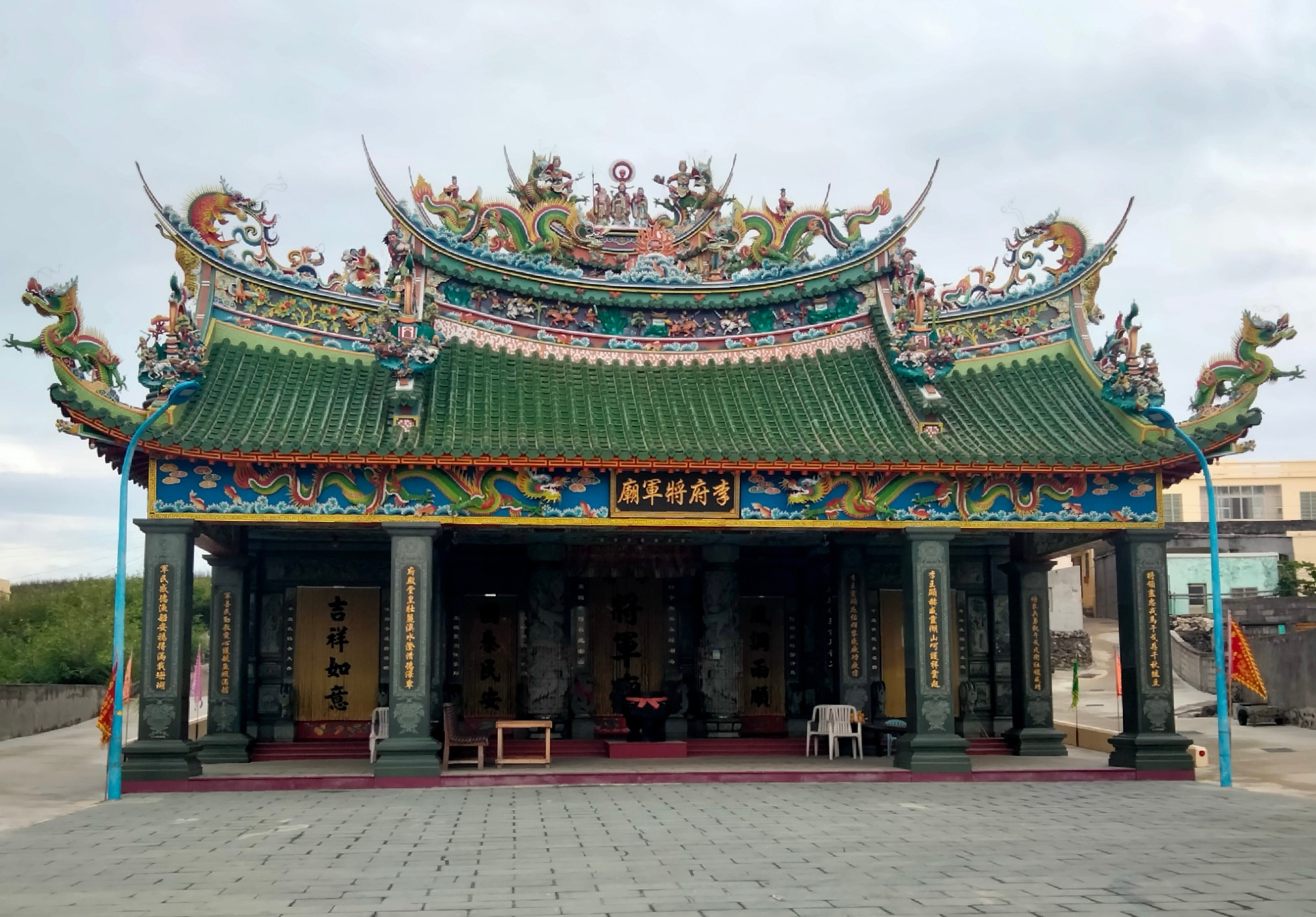 一座傳統中式廟宇正立面，綠色琉璃瓦屋頂，兩端翹角飾有金色龍雕，屋檐下方有藍色木質裝飾和金色文字牌匾，四根綠色石柱支撐寬敞的廟堂，前方為紅色平台和淡藍色支架。