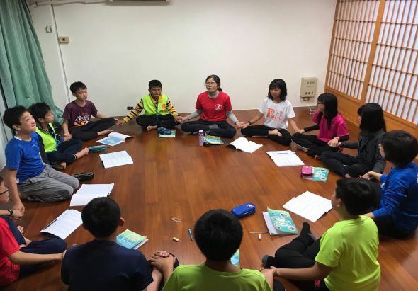 約十五名青少年圍坐在木製長桌周圍進行小組活動。參與者穿著各色上衣，桌上放有藍色書籍和白色文件。室內有綠色窗簾和木質隔屏，呈現教室或活動室的環境。