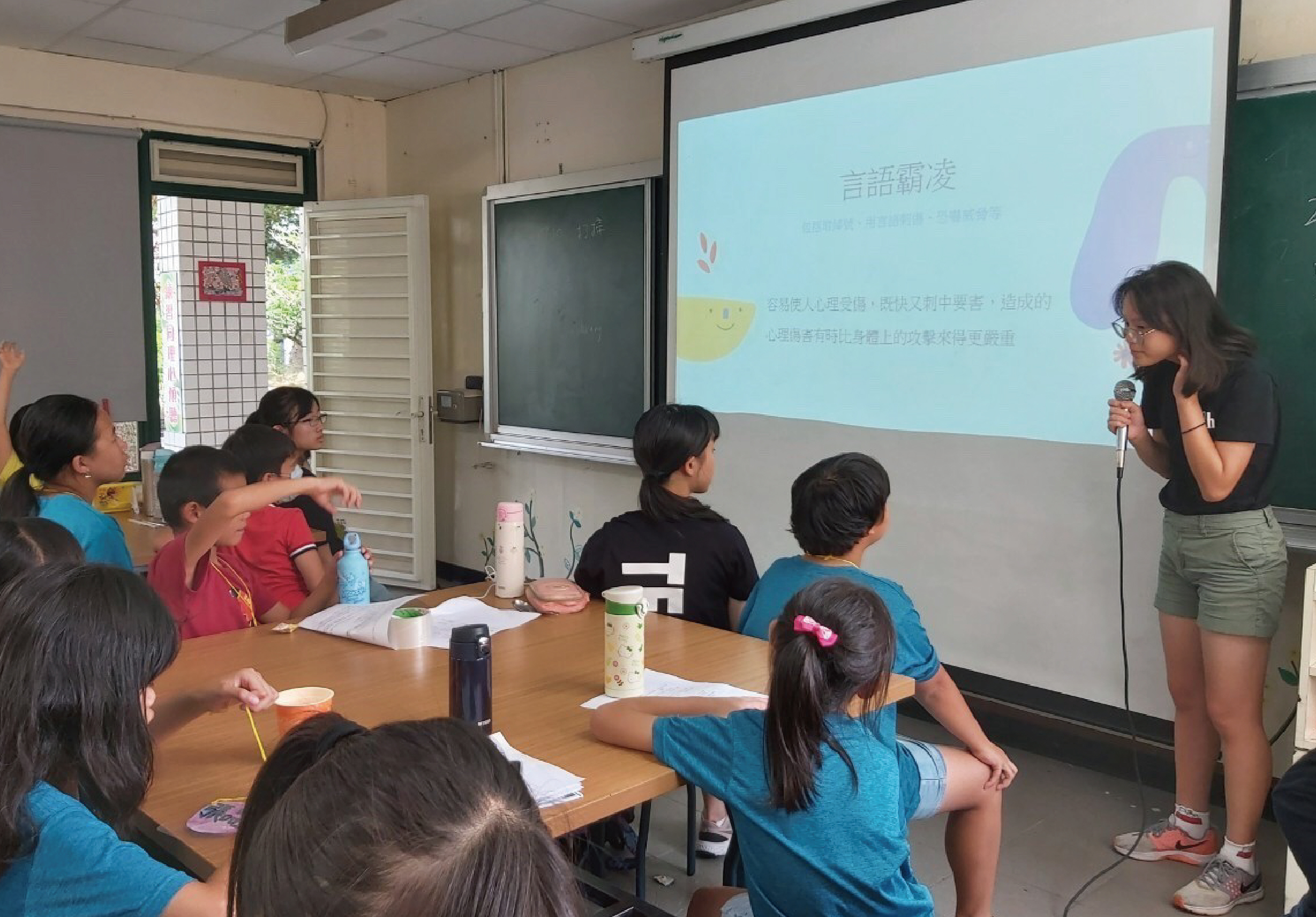課堂上，一位女性講者手持麥克風向學生們講解。投影幕上展示著繁體中文內容「言語涵養」。多位穿著藍色和紅色制服的小學生坐在木桌前，認真聆聽和觀看投影內容。教室裡有黑板、門窗和天花板投影機等設施。