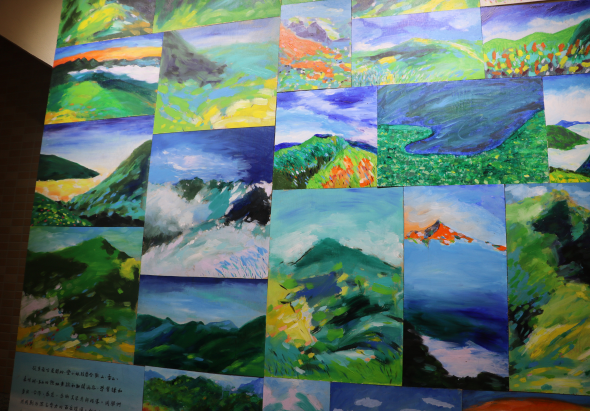美術展覽牆面展示約二十五幅風景油畫作品，以山水、湖泊、田野為主題，運用鮮豔的綠色、藍色、黃色等色調，呈現四季山林景觀。作品排列成網格狀，左下方有展覽說明文字牌。