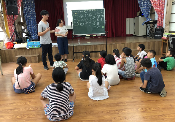 教室內，一名男性教師和一名女性助教站在講台上，面向圍坐成圈的約十五名小學生。講台後方有綠色黑板和投影幕。學生們認真聆聽課程內容，教室布置有紅藍色窗簾和教學用具。