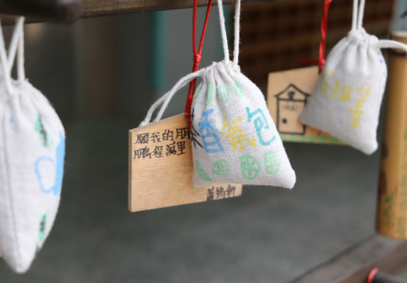 數個白色布製小袋子懸掛在木製架子上，袋身印有彩色字母和中文文字，附紅色或白色繩子。木製標籤上亦有中文文字。背景為模糊的室內環境，呈現手工藝品或教學成果展示的場景。
