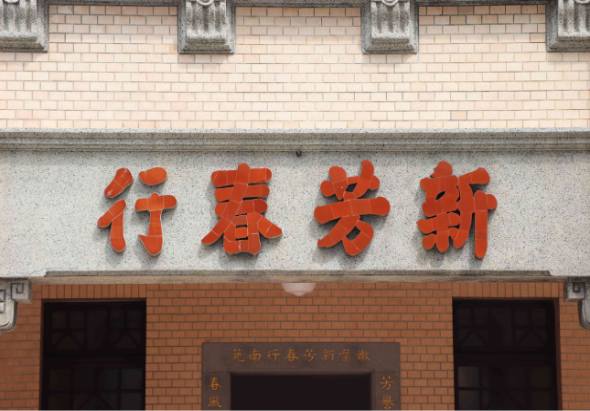 磚紅色建築物外牆，掛著紅色立體字招牌「新芳春行」，上方為米色磚砌牆面，下方為紅磚牆面，中央有兩個深色門窗，門上方有褐色牌匾，建築具有古典商業建築風格。