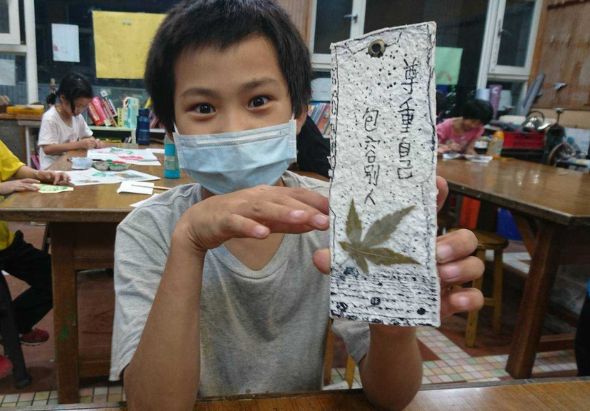 教室內，戴著口罩的小男孩展示自己製作的白色手工藝品，上面有日文書寫和綠色植物裝飾。身後同學正進行美術創作活動。
