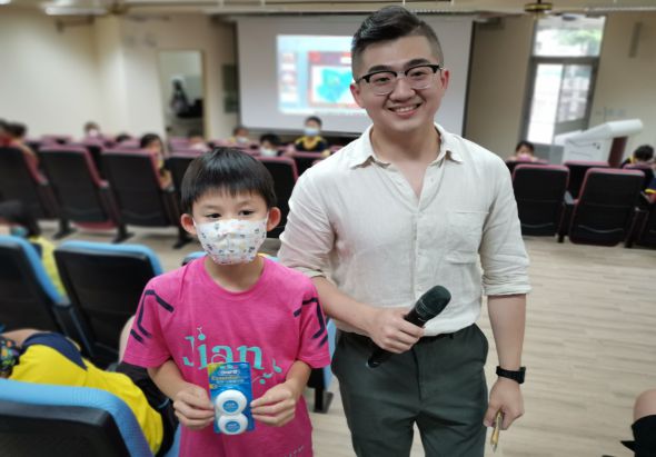 一名戴眼鏡的年輕男性和一名穿粉紅色衣服戴口罩的年幼男童在室內活動場地合照。男性穿著白色襯衫手持麥克風，背景有投影螢幕和紅色座椅。