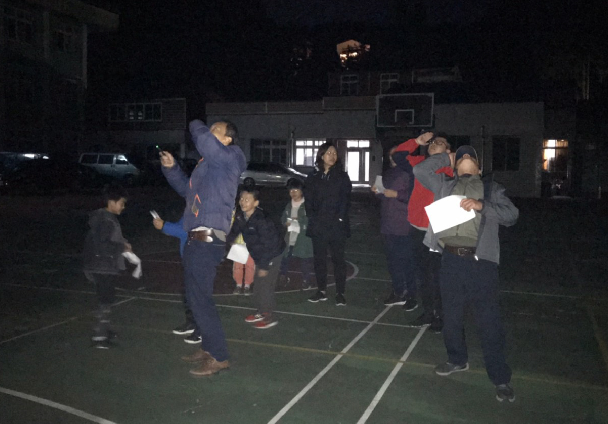 夜間籃球場上，兩位成人正帶領一群兒童進行投籃練習。左側一人舉起手臂做投籃動作，右側一人扶著兒童協助投籃。約十名兒童穿著冬季衣物圍觀學習。背景為學校建築和停泊的車輛。