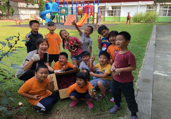 一群小學生與女老師在校園草坪上聚集，手持著一個紙箱，開心地擺出各種姿勢拍照。背景可見彩色遊樂設施、校舍與綠地。學生們穿著橙色及紅色衣服，氣氛歡樂溫馨。