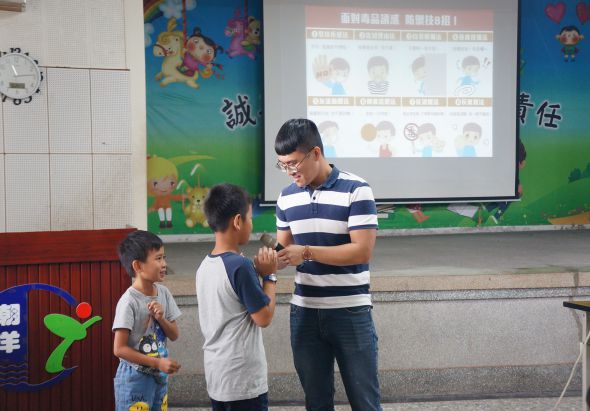 教室內，一位穿著藍白條紋衣服的男性教師與兩位小學生互動，背後投影幕上顯示兒童衛生健康教育內容，包含多個卡通人物頭像，牆上裝飾有彩色兒童圖案。
