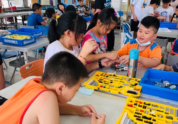 小學生在教室內進行STEM教育活動，穿著橙色背心的兒童圍坐在桌邊，操作黃色零件工具箱進行科學實驗或機械組裝，老師和同學在一旁指導協助。