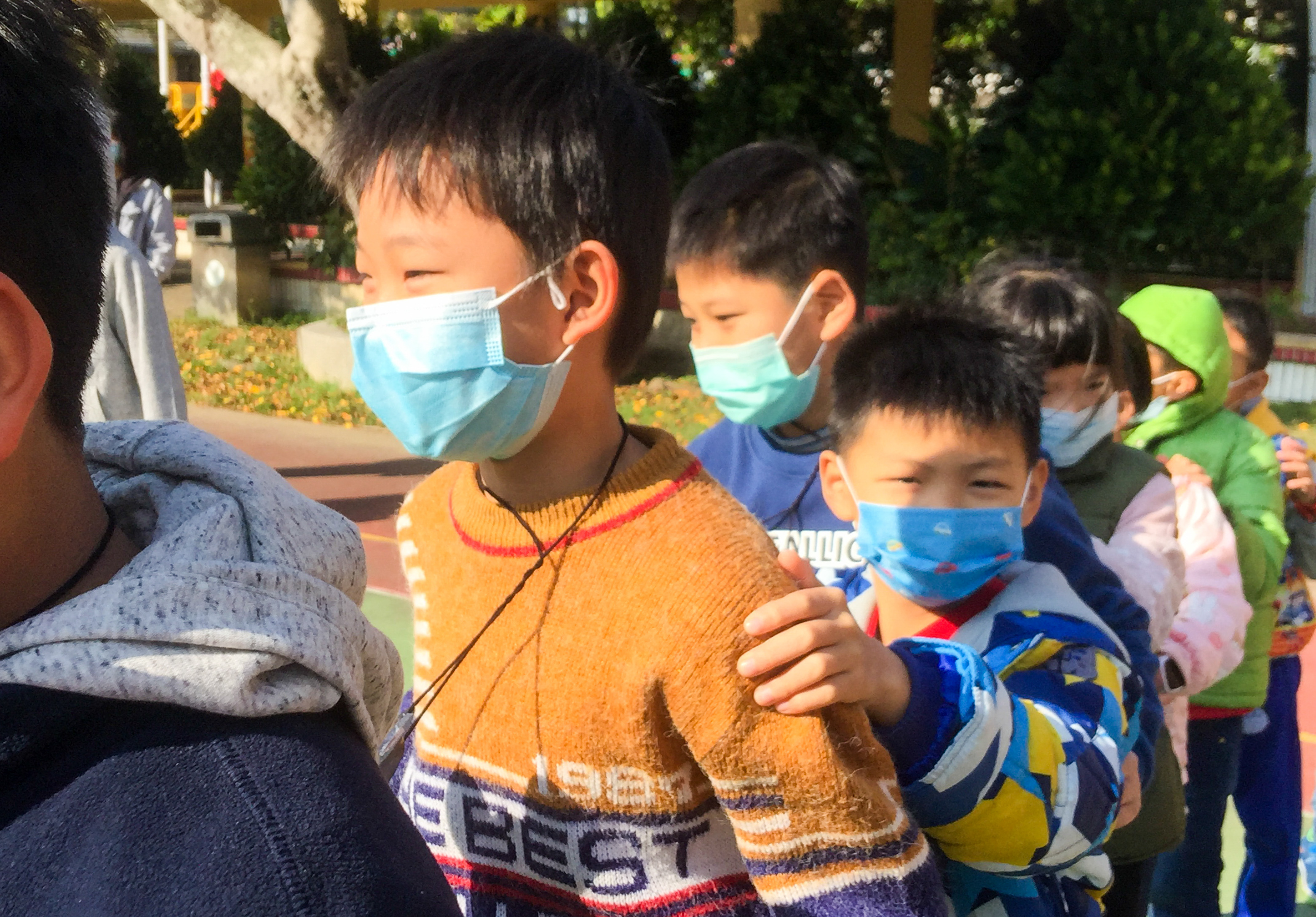 一群年幼的小朋友在戶外遊樂場活動，多數人配戴口罩。前景中穿著黃色毛衣的男童與穿著藍色衣服的男童緊靠在一起，後方還有其他穿著彩色衣服的孩童。背景可見綠樹和建築物，陽光灑落在遊樂場上。