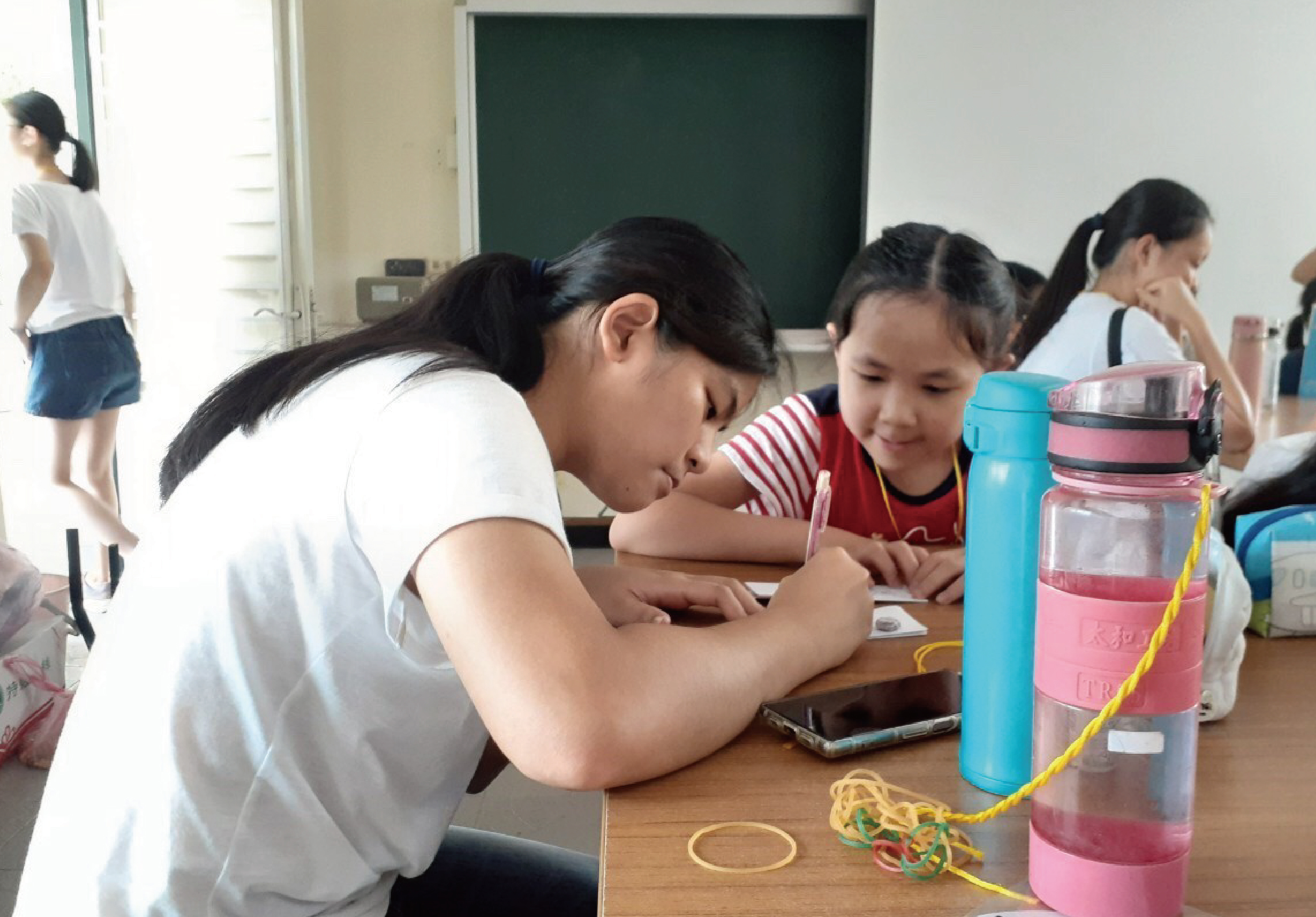 教室內，一位老師和多名學生在桌前進行科學實驗活動。學生們觀察著裝有粉紅色液體的容器和黃色繩索，專注地進行探究學習。背景可見黑板和其他學生。