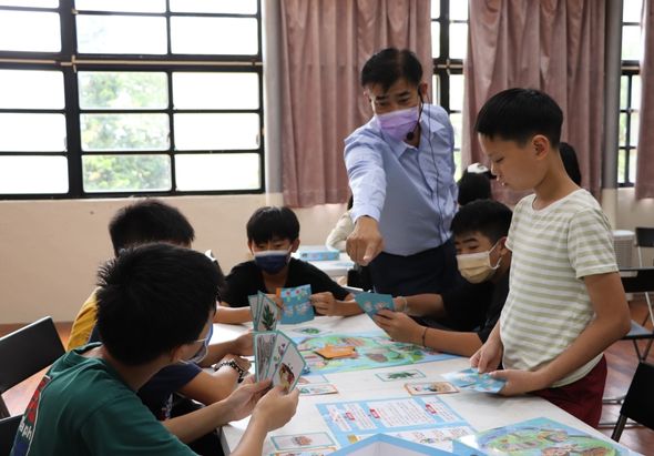 一位戴著口罩的男性教師在教室裡向坐著的六名小學生講授課程。學生們圍坐成一圈，桌上放著教材和圖書。教室背景有大窗戶和褐色窗簾。
