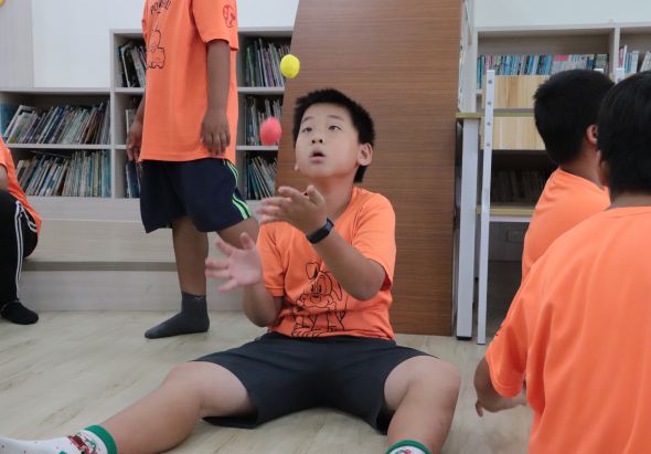 在圖書館內，穿著橙色上衣的小男孩坐在地上，抬頭看著空中的黃球，雙手做出接球的姿勢。其他穿著相同橙色上衣的小孩站立在旁邊觀看，背景是書架。