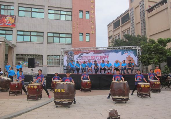 學校操場上舉辦文藝表演活動，舞台後方掛著藍紅色布幔，台上坐著穿著藍色制服的學生，台前排列多面大鼓，表演者穿著紫色服裝正進行鼓樂演奏，背景是校園建築物。