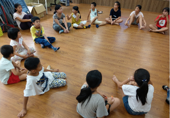 一群成人和兒童坐在木質地板上圍成圓圈進行互動活動，孩子們在中央玩耍，成人站在後方觀看。室內環境明亮，牆面為木製裝潢，呈現溫暖的教室或社區活動空間氛圍。
