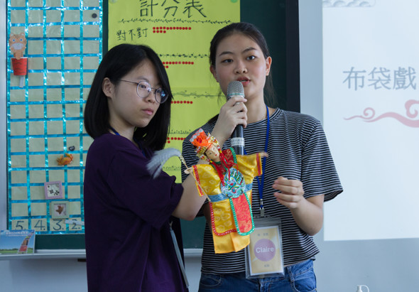 兩名學生在教室進行活動展示，左邊學生穿著深紫色衣服，右邊學生穿著條紋T恤持麥克風，手持彩色布袋道具。身後黑板上有中文教學內容，牆上貼有教學海報和計畫表。