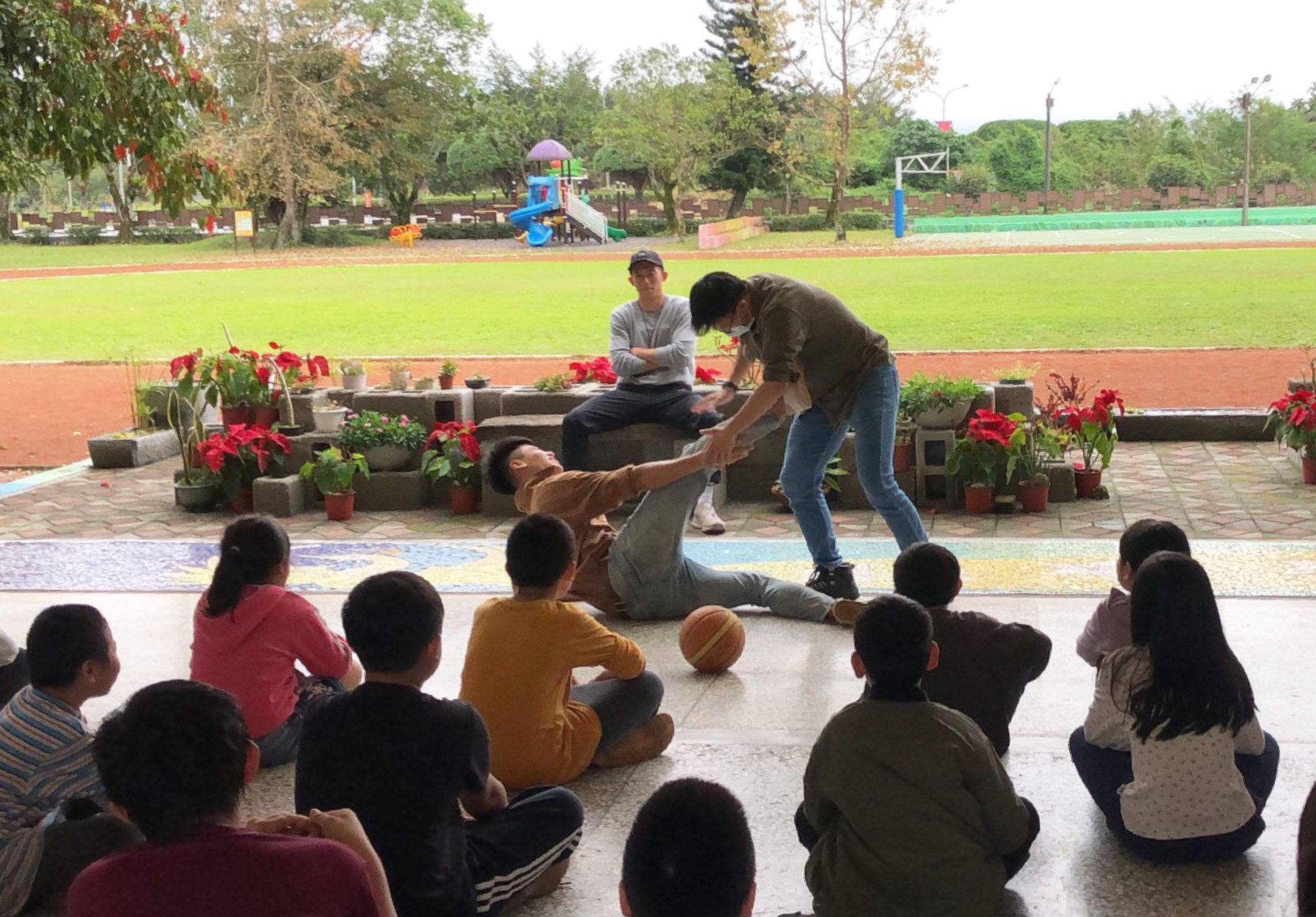 公園內進行戶外教學活動，一位教師在眾多坐地上的小學生面前示範籃球動作，背景為運動場、花圃和遊樂設施，另有成人在旁協助。