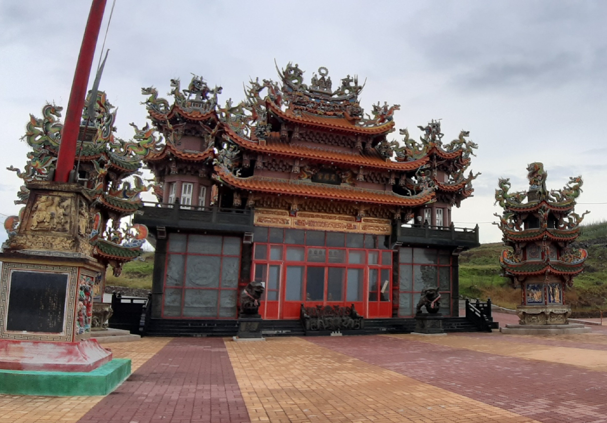 壯觀的中式廟宇建築，具有多層飛簷、金色裝飾和綠色龍雕。紅色大門和窗戶為主體，兩側配置精美的塔樓。廣場鋪設紅磚，左側立有紀念碑。整體呈現傳統東方宗教建築的莊嚴風貌。