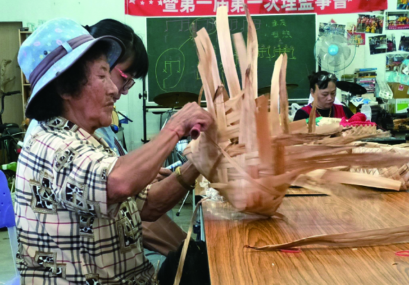 木工課堂上，戴著藍色帽子的男性講師正向學生展示一件木製手形雕刻作品。教室後方有綠色黑板、紅色標語布條和教學用具。學生坐在木製工作台旁認真觀看示範。