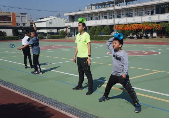學校操場上，數名學生進行羽毛球運動。穿著螢光黃背心的教練在中央指導，學生們頭戴羽毛球拍握把，練習接球動作。背景為校舍建築和花卉裝飾。