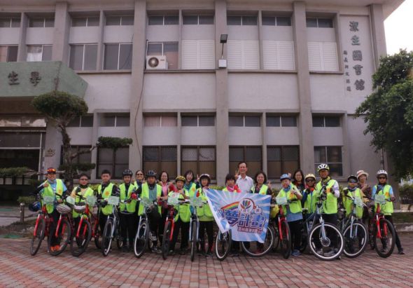 一群穿著螢光黃綠背心的人員，騎著自行車聚集在學校建築前的紅磚廣場上，中央舉著藍色旗幟，參與單車運動推廣活動。