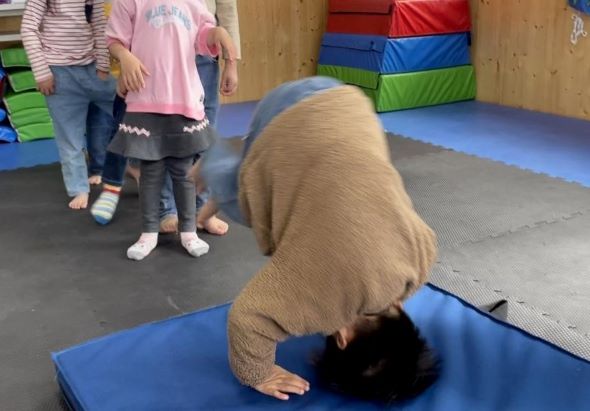 一名穿著棕色毛衣的孩童在藍色墊子上做倒立動作，展示體操技能。背景有其他兒童站立觀看，室內設有彩色軟墊和木製牆面，呈現體操或運動課程的場景。