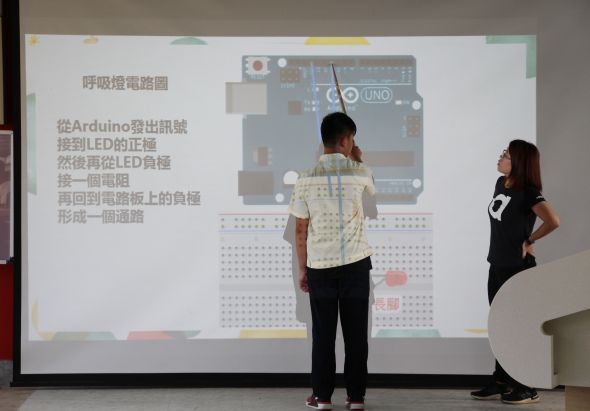 兩名學生在教室進行Arduino電路實驗教學演示。投影幕上展示呼吸燈電路圖與步驟說明，包括Arduino輸出腳位、LED正負極接線及電阻配置等內容。