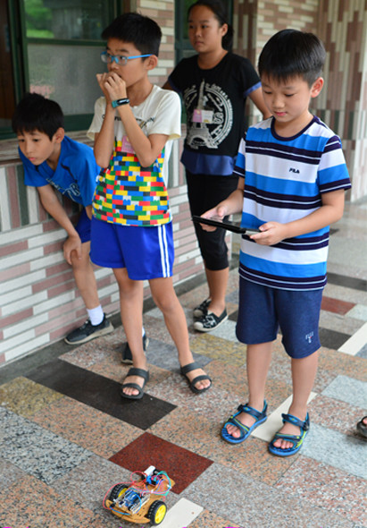 四名男孩在室內走廊進行遙控車競賽，其中三人穿著藍色運動服，一名大人在旁監督。前方地面上放置黃藍色遙控小車，孩子們專注操控比賽進行。