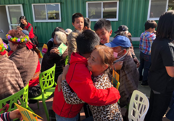 綠色鐵皮建築前，多位大人和小孩聚集參加戶外活動。人們穿著五顏六色的衣服，有人抱著孩子，有人手持花束，彼此親切互動。白色塑膠椅散落周圍，背景可見多扇窗戶。