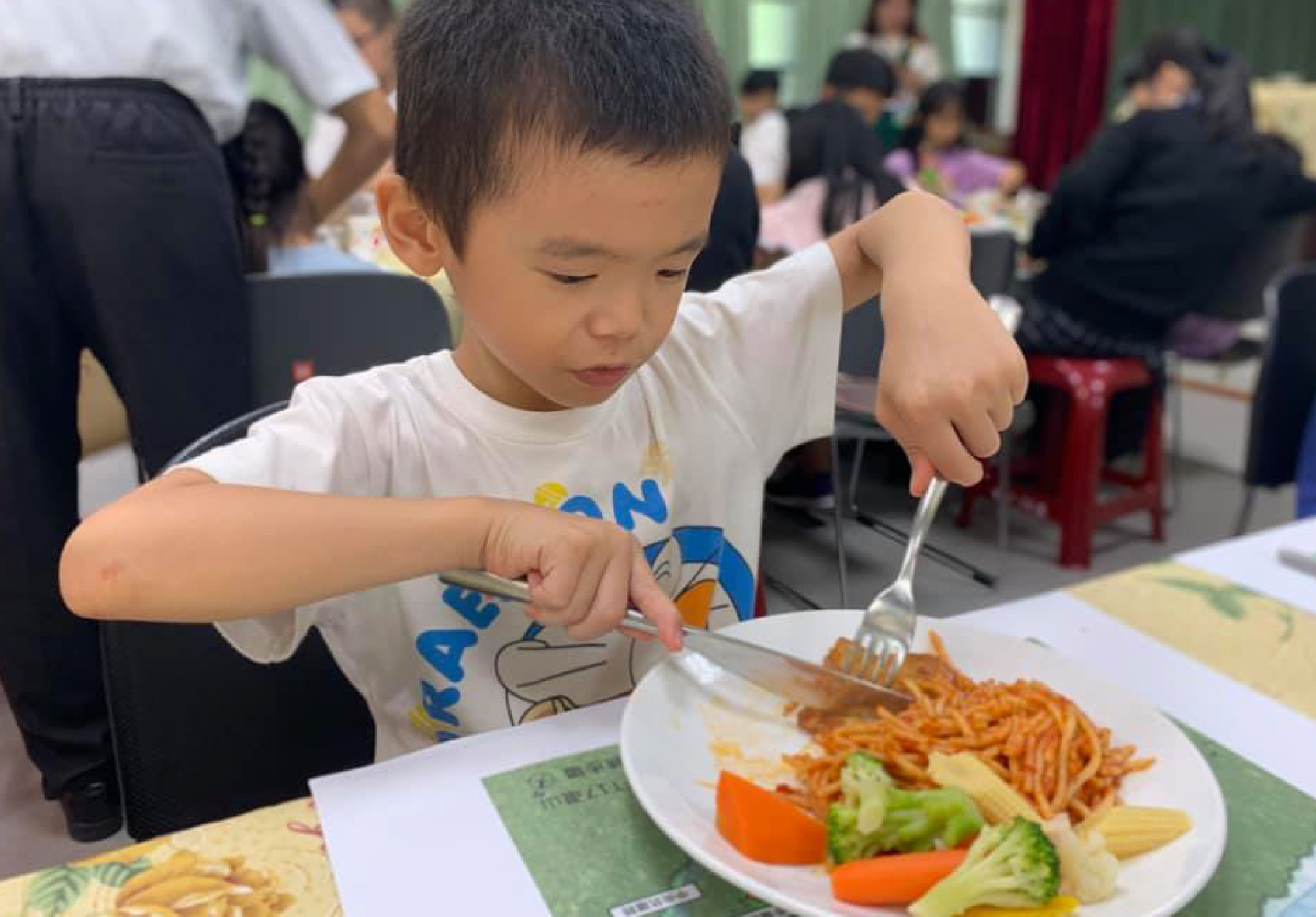 小男孩在學校食堂進行用餐活動，穿著白色卡通T恤，專注地用叉子切著盤中的炸薯條。盤子裡還有紅蘿蔔、綠色蔬菜和黃色薯條。背景可見其他學生和老師進行同樣的餐飲活動。