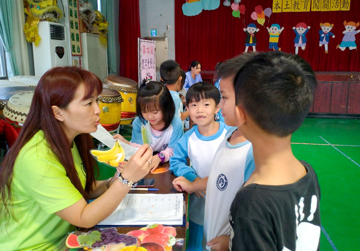 幼稚園教室內，穿著螢光綠衣服的女性教師手持香蕉和水果圖卡，向圍成一圈的小朋友們進行營養教育。背景有紅色布景和彩色兒童貼紙裝飾。
