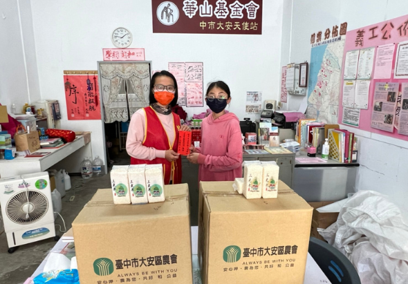 兩名戴著口罩的女性在辦公室內站在兩個臺中市政府農業處的紙箱前合照。背景牆上貼著紅色春聯、粉色公告欄和各式海報。左側放置空調、右側有書架和辦公用品。
