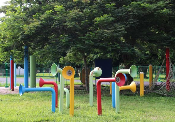 公園內的彩色音樂遊樂設施，由多根藍、紅、黃、綠色管狀結構組成，頂端裝有黃色、紅色等敲擊樂器。綠樹成蔭的背景中可見兒童遊樂場設施，草地上擺放著互動音樂裝置。