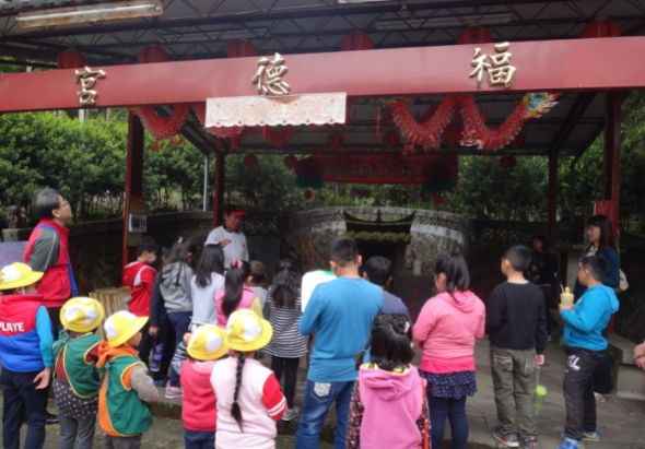 一群小學生和家長在中式廟宇前排隊參訪，紅色建築上懸掛著「宮德福」字樣和紅色裝飾，部分遊客配戴黃色安全帽，進行校外教學或導覽活動。