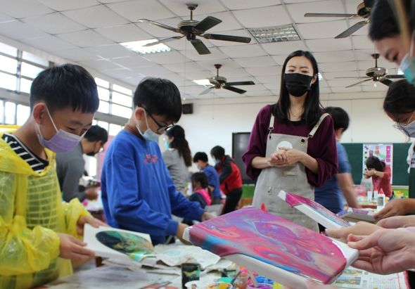 室內藝術工作坊，多名戴口罩的學生圍著桌子，在老師指導下進行繪畫創作。學生們手持畫筆，在色彩繽紛的畫板上作畫，天花板裝有風扇和燈具，窗戶透進自然光。