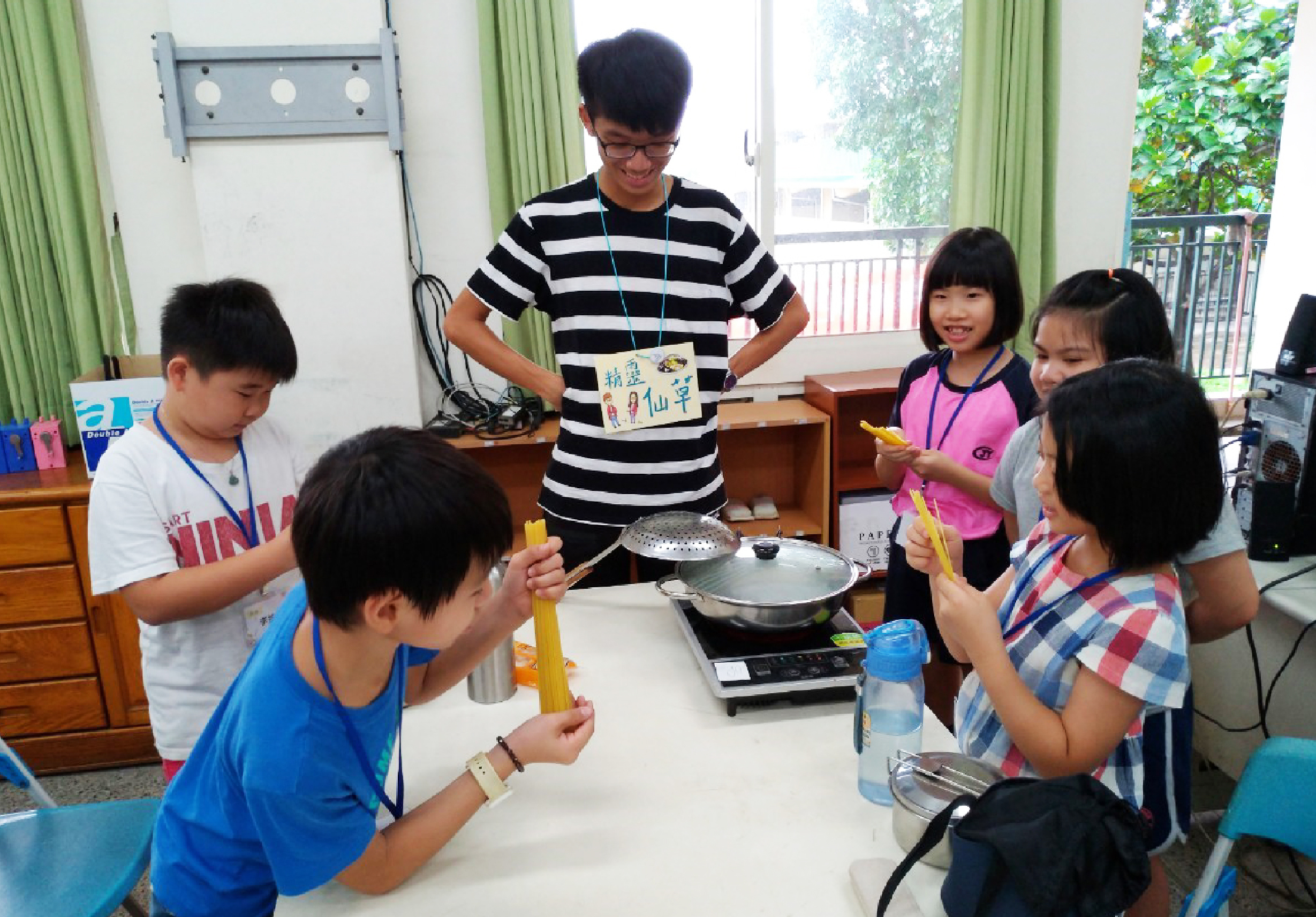 一位男性老師在教室裡向六位學生示範烹飪，桌上放著鍋具和食材。學生們圍成一圈，手持廚具，認真觀看老師的示範。教室明亮寬敞，窗外可見綠色植物。這是一場實踐性的烹飪教學活動。