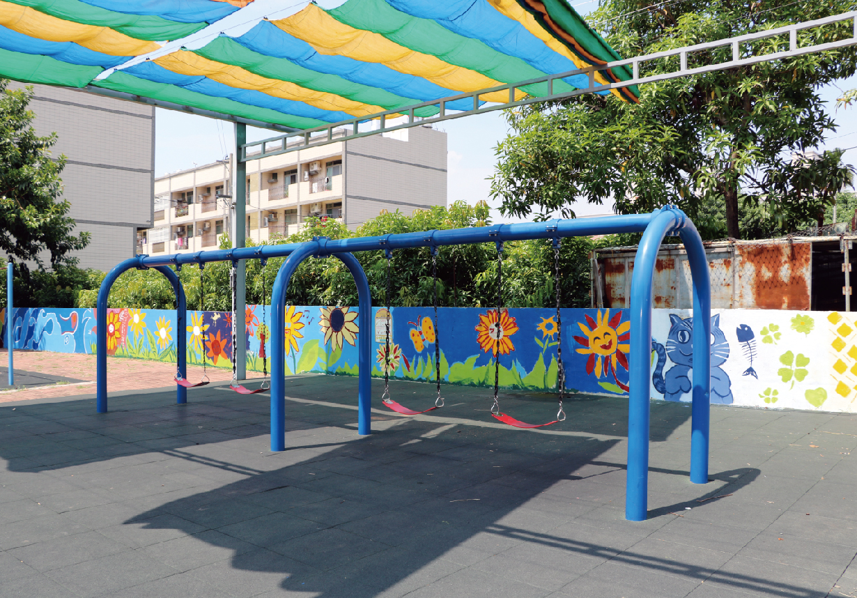 幼兒園遊樂場，藍色鞦韆架配彩色布篷頂，背景為教學樓。欄杆繪有向日葵、太陽和動物圖案，黑色橡膠地面增強安全性。綠樹環繞，陽光充足，適合兒童遊玩的戶外活動空間。
