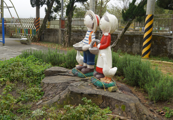 公園內的卡通人物雕塑，兩隻白色兔子角色穿著藍紅色衣服，站在石頭上釣魚。周圍綠植環繞，遠處可見柵欄和遊樂設施，為兒童公園場景。