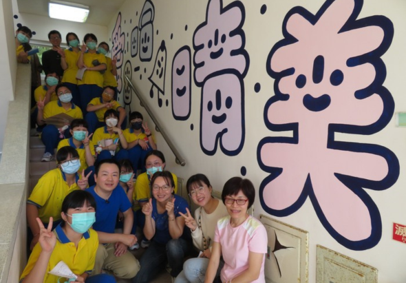 一群穿著黃色制服的學童與五位成人在室內合照，背景牆上有藍色和粉紅色的卡通人物插畫和中文字。大多數人戴著口罩，擺出開心的姿勢，看起來像是學校或教育機構的集體活動或慶祝活動。