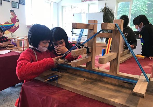 幼兒園教室內，穿著紅色毛衣的小男孩和女孩一起操作木製軌道玩具，專注地推動小車沿著藍色軌道滑行。背景有其他孩童在玩耍，室內鋪設紅色地墊，窗戶採光良好。